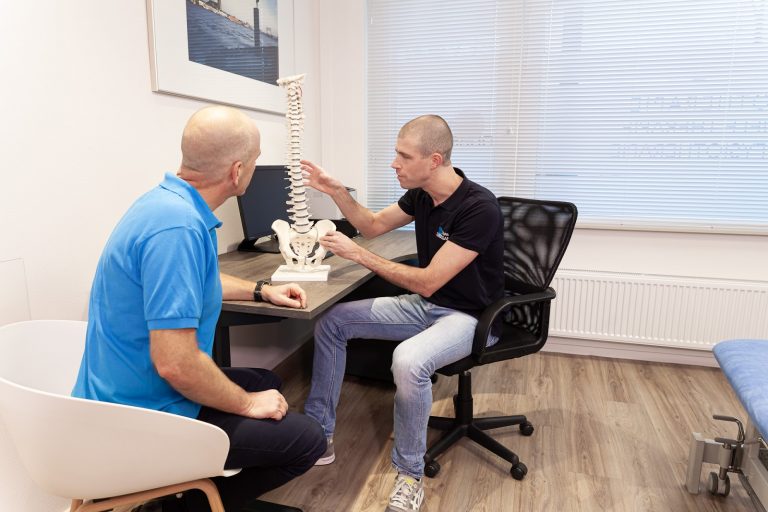 Fysiotherapeut Joost in het zwarte shirt, legt uit welke rugklachten patiënt Peter in het blauw precies heeft door middel van een prop