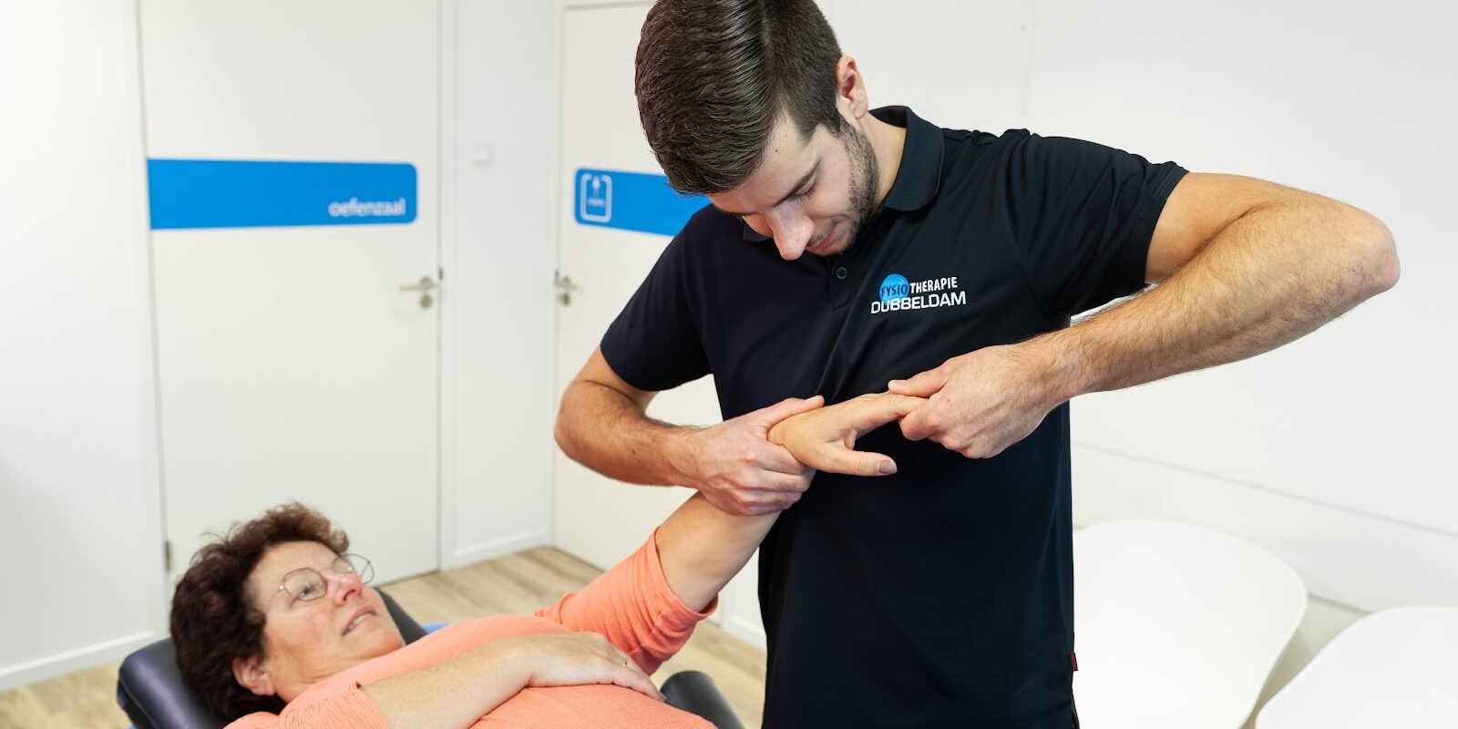 Fysiotherapeut Reno Bosman in een zwart shirt helpt een vrouw in een zalmroze shirt op de behandelbank aan haar hand door middel van hand- en polstherapie