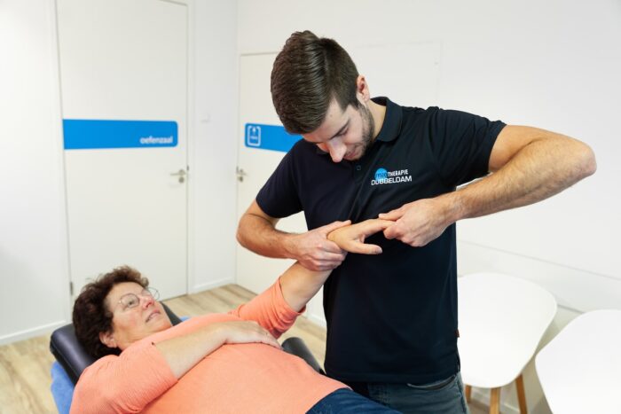 Hand- en polstherapie Fysiotherapeut Reno Bosman in een zwart shirt helpt een vrouw in een zalmroze shirt op de behandelbank aan haar hand door middel van hand- en polstherapie