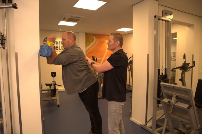 Een fysiotherapeut en een patiënt staan in een oefenzaal. De patiënt steunt tegen de muur, terwijl de fysiotherapeut zijn handen op de rug van de patiënt met aspecifieke rugklachten houdt.