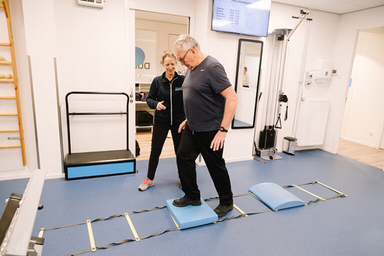 Fysiotherapeut staat naast een man in een zaal. Man doet aan dagelijks bewegen. Hij loopt op een looptrap voor oedeemtherapie