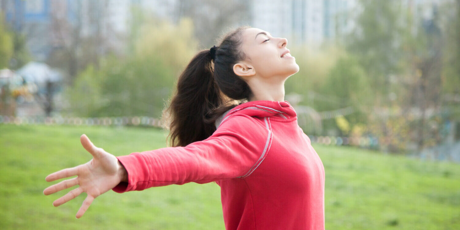 Een vrouw met een rode trui aan, staat op een grasveld. Ze heeft haar armen uitgestrekt. Ze oefent met zang- en ademhalingstechnieken.