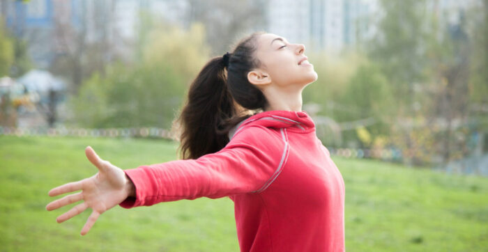 Zang- en ademhalingstechnieken Een vrouw met een rode trui aan, staat op een grasveld. Ze heeft haar armen uitgestrekt. Ze oefent met zang- en ademhalingstechnieken.