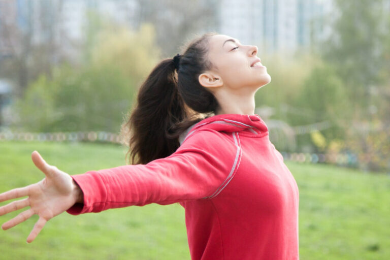 Een vrouw met een rode trui aan, staat op een grasveld. Ze heeft haar armen uitgestrekt. Ze oefent met zang- en ademhalingstechnieken.