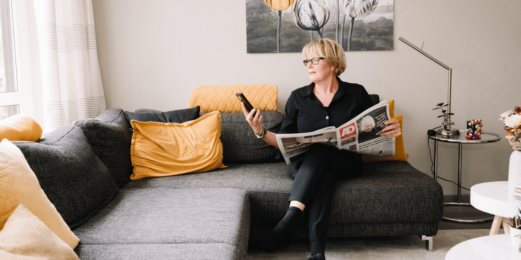 Vrouw heeft klachten tijdens ontspanning en kijkt op haar telefoon op de bank terwijl zij een krant leest in de woonkamer